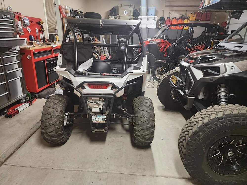 UTV with oversized tires and racing seats in a garage, surrounded by tools and other off-road vehicles.